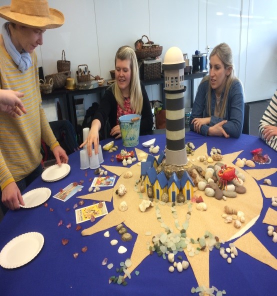 Image from a Dementia Champion training day - four people gathered around a table with different objects including a model lighthouse, sea shells, paper plates, cups and stones