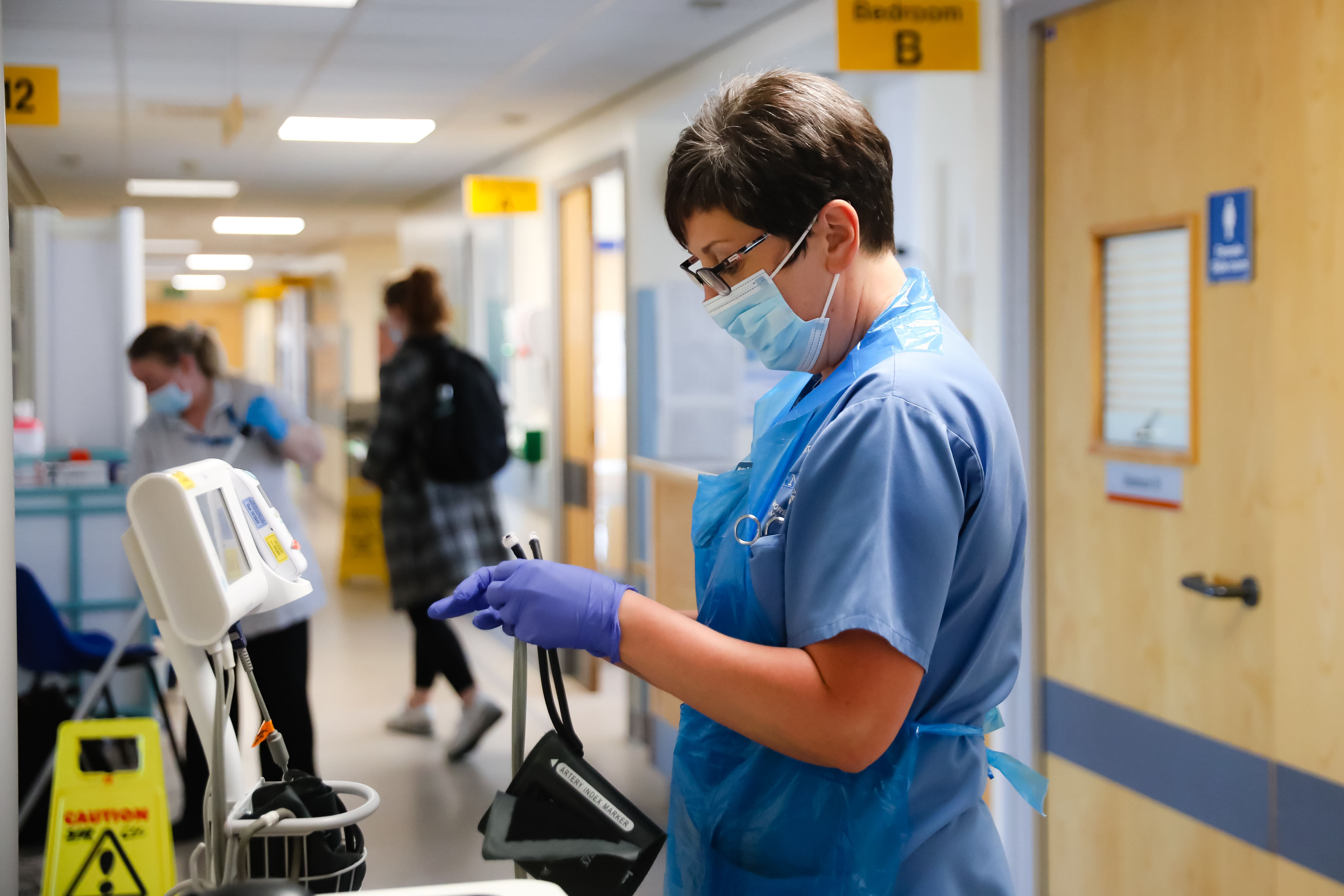 A Morecambe Bay NHS staff member in a blue uniform stood in a corridor with a face mask and gloves on holding a blood pressure cuff