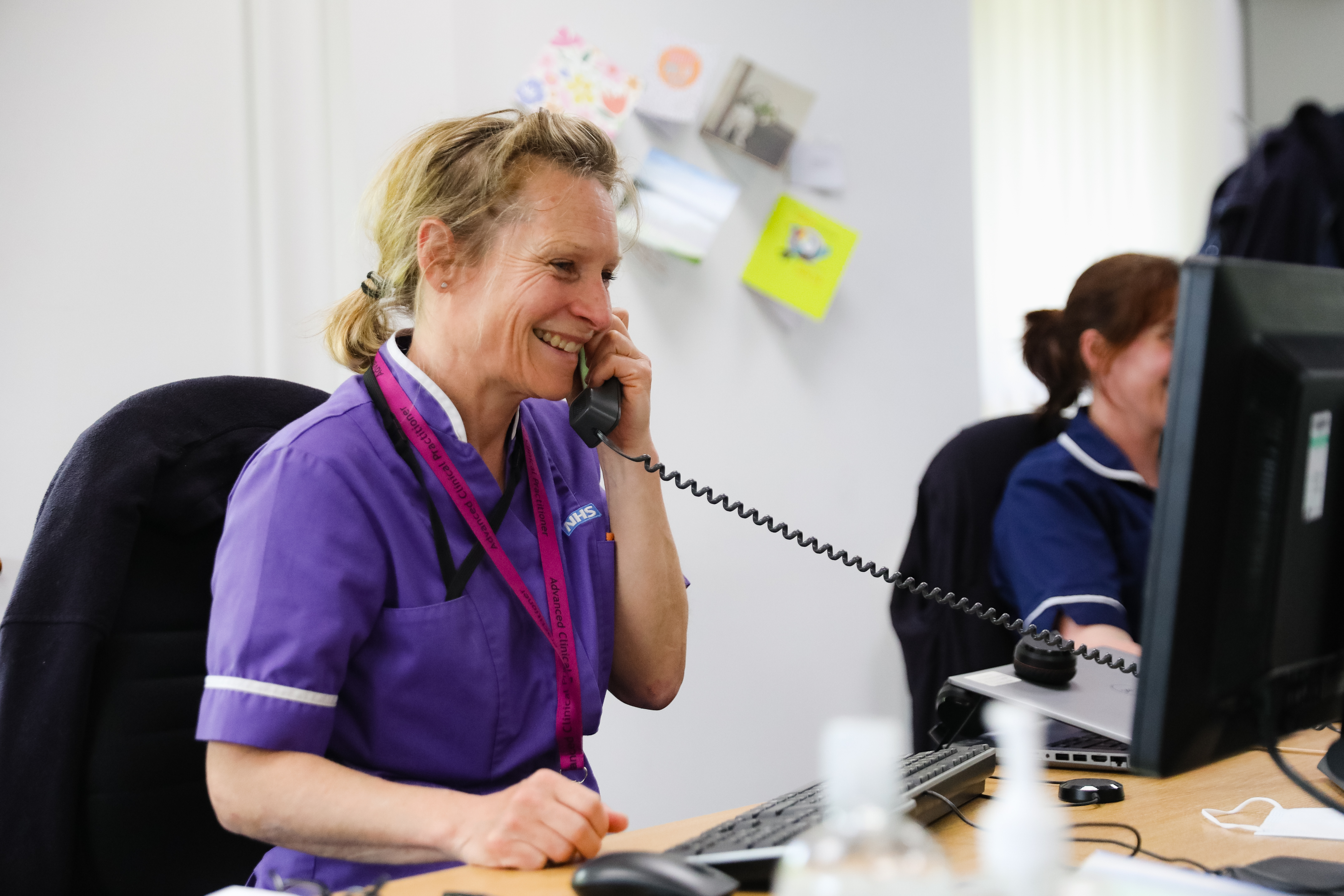 A Morecambe Bay NHS colleague in purple uniform sitting at a desk and smiling while on the phone