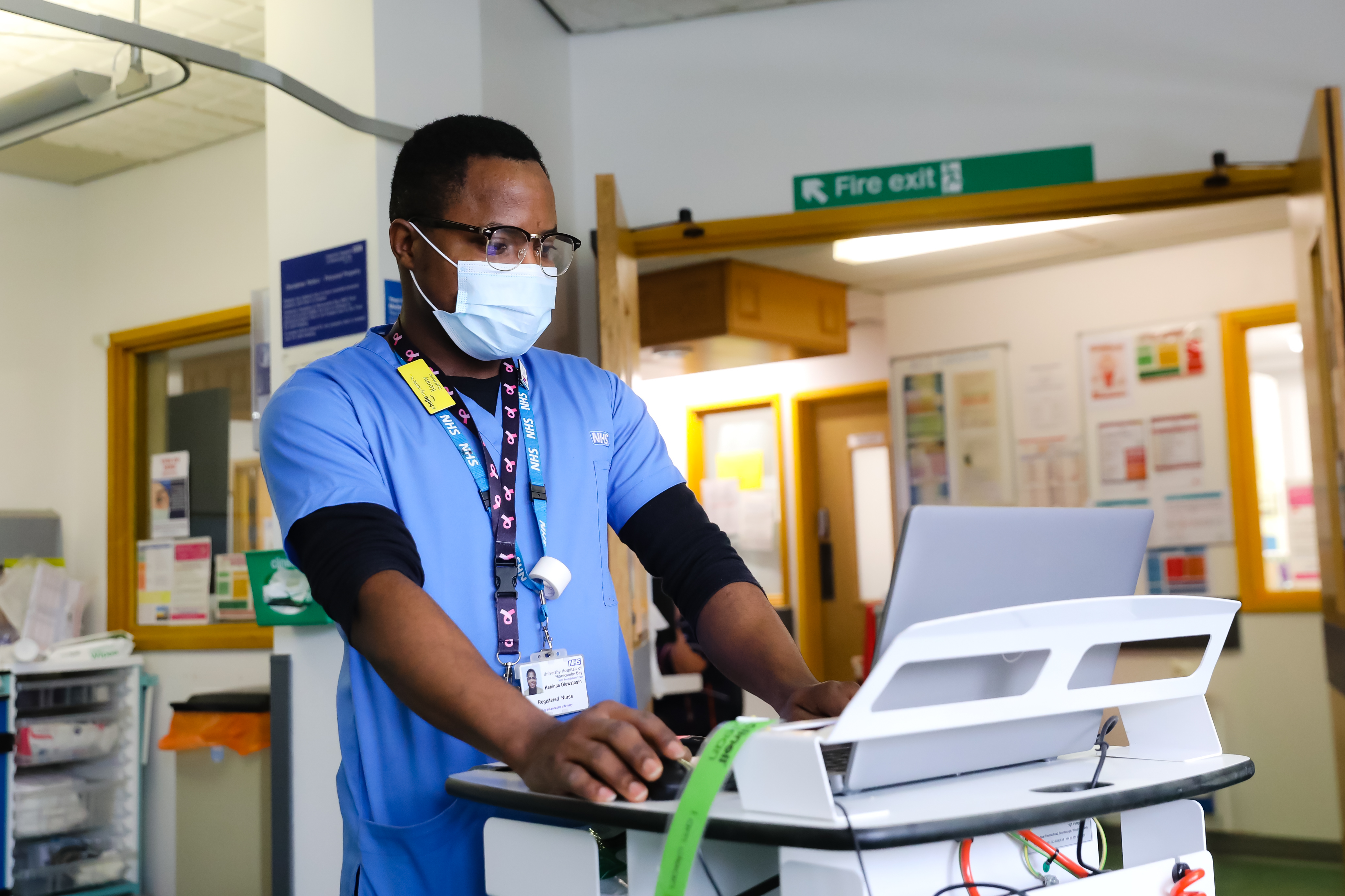 A Morecambe Bay NHS Nurse in blue scrubs standing and using a laptop on a trolley