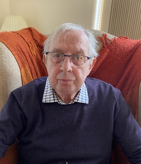 An elderly man with glasses and a navy jumper sitting on a brown patterned sofa and looking directly at the camera.