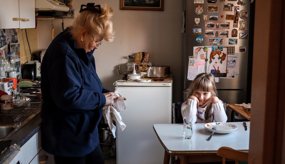 A grandmother talks to her granddaughter in a small kitchen. A bowl of unfinished food sits on the table, and washing up is visible in the background. A fridge with magnets and drawings is visible in the background. 