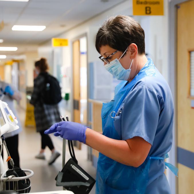 A Morecambe Bay NHS staff member in a blue uniform stood in a corridor with a face mask and gloves on holding a blood pressure cuff